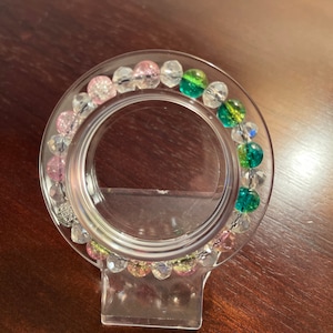 Puede incluir: Un brazalete transparente con cuentas rosas, verdes y transparentes. El brazalete se muestra en un soporte de plástico transparente.
