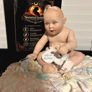 May include: A porcelain baby doll with light skin, pinkish eyes, and a small tuft of blonde hair tied with a pink ribbon. The doll holds a white fabric tag labelled SPECIAL DELIVERY and wears a matching tag. It sits on a quilted pastel blanket.