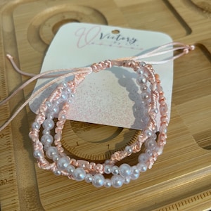 Op de afbeelding: Een roze en witte kralen armband met een roze touwtje eromheen gebonden. De armband ligt op een houten oppervlak met een liniaal en een label met de tekst "Victory".