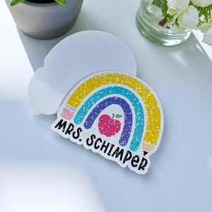 May include: A colorful rainbow-shaped sticker with glitter accents, featuring a red apple and the text "MRS. SCHIMPER". The sticker is on a white, cloud-shaped base. The design includes yellow, blue, and pink colors.