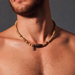 Peut inclure: Un collier de perles composé de perles en bois brun clair, de petites entretoises métalliques et d'une perle centrale cylindrique brun foncé à motifs. Une petite breloque métallique est suspendue au centre du collier, muni d'un fermoir.