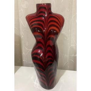 May include: A decorative vase in the shape of a female torso. The vase is primarily red with a swirling pattern of black stripes. The vase has a glossy finish and is displayed on a white surface.