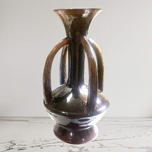 May include: A brown ceramic vase with a glossy finish. The vase has a bulbous body, a flared neck, and four curved handles. The base is a darker brown, and the interior is white.
