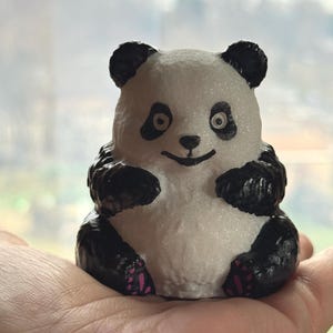 May include: A small, handcrafted panda figurine with a white body and black markings on its ears, arms, and legs. The panda has a smiling face with black eyes and a pink accent on its feet. The figurine is a cute, decorative item.