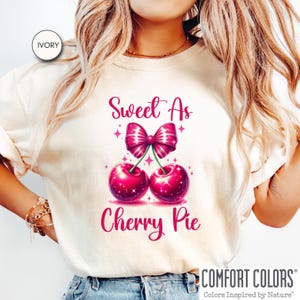 May include: A white t-shirt with the text "Sweet As Cherry Pie" in pink lettering. The text is accompanied by a graphic of two pink cherries with a pink bow on top.