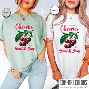 May include: Two women wearing t-shirts with a graphic of cherries on a branch. The text on the shirts reads "Fresh Picked Cherries 100% Organic Sweet & Juicy". The shirts are available in bay and white.
