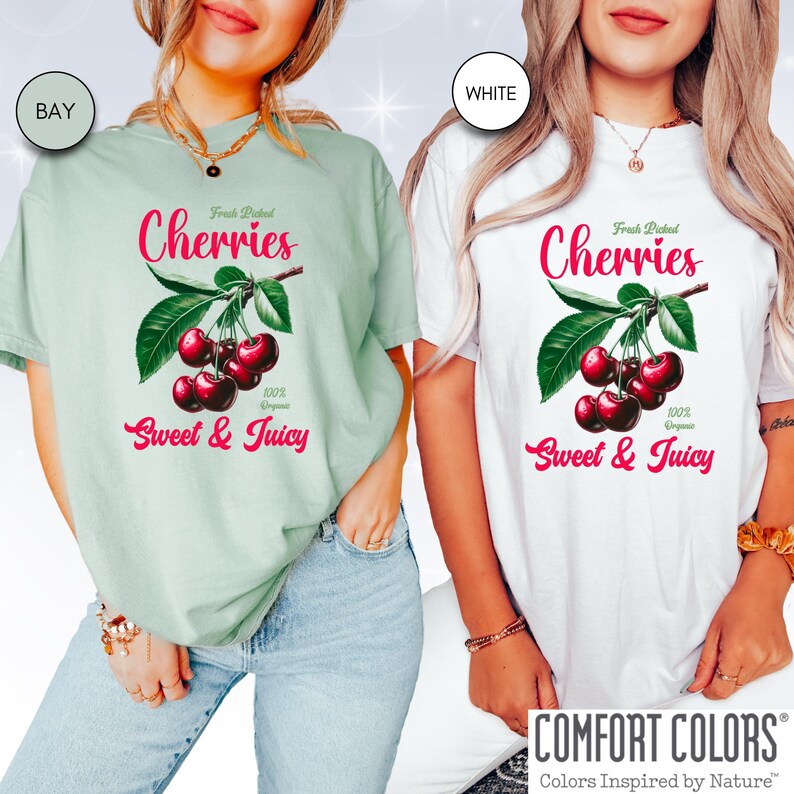 May include: Two women wearing t-shirts with a graphic of cherries on a branch. The text on the shirts reads "Fresh Picked Cherries 100% Organic Sweet & Juicy". The shirts are available in bay and white.