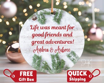 May include: A round ceramic ornament featuring a white wood grain background with green pine branches and pinecones. The text reads: Life was meant for good friends and great adventures! Andrea & Aubree. It hangs from a gold cord.