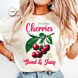 May include: A white t-shirt with a graphic of red cherries on a branch with green leaves. The text "Fresh Picked Cherries" is above the cherries and "100% Organic Sweet & Juicy" is below the cherries.