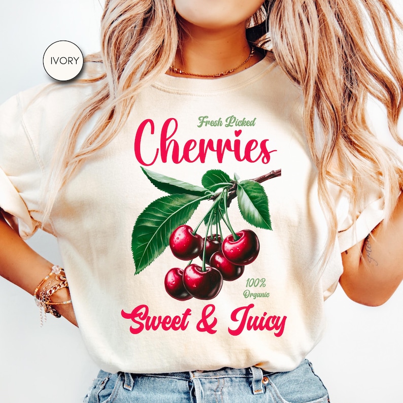 May include: A white t-shirt with a graphic of red cherries on a branch with green leaves. The text "Fresh Picked Cherries" is above the cherries and "100% Organic Sweet & Juicy" is below the cherries.