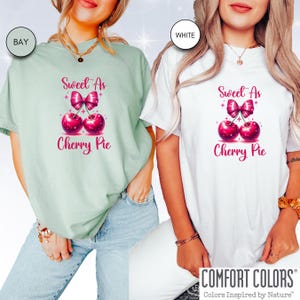 May include: Two t-shirts with a graphic of two cherries with a pink bow and the text "Sweet As Cherry Pie". The t-shirts are in the colors bay and white.