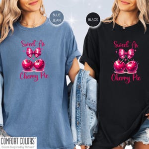 May include: Two women wearing t-shirts with the text "Sweet As Cherry Pie" in pink lettering. The shirts feature a pink cherry with a pink bow on top. One shirt is blue jean and the other is black.