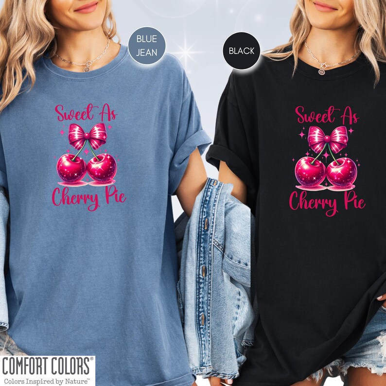 May include: Two women wearing t-shirts with the text "Sweet As Cherry Pie" in pink lettering. The shirts feature a pink cherry with a pink bow on top. One shirt is blue jean and the other is black.
