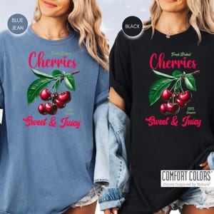 May include: Two t-shirts with a graphic of red cherries on a branch with green leaves. The text "Fresh Picked Cherries" and "Sweet & Juicy" is printed on the shirts. The shirts are available in blue jean and black.