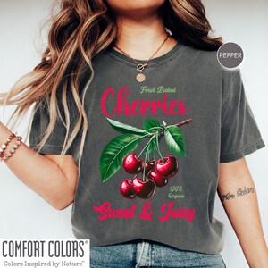 May include: A gray t-shirt with a graphic of red cherries on a green stem with leaves. The text "Fresh Picked Cherries" is above the cherries and "Sweet & Juicy" is below the cherries. The text "100% Organic" is to the right of the cherries.