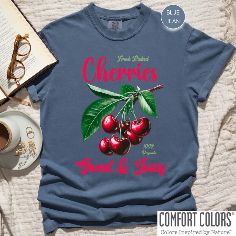 May include: A blue jean t-shirt with a graphic of red cherries on a branch with green leaves. The text "Fresh Picked Cherries 100% Organic Sweet & Juicy" is printed on the shirt.