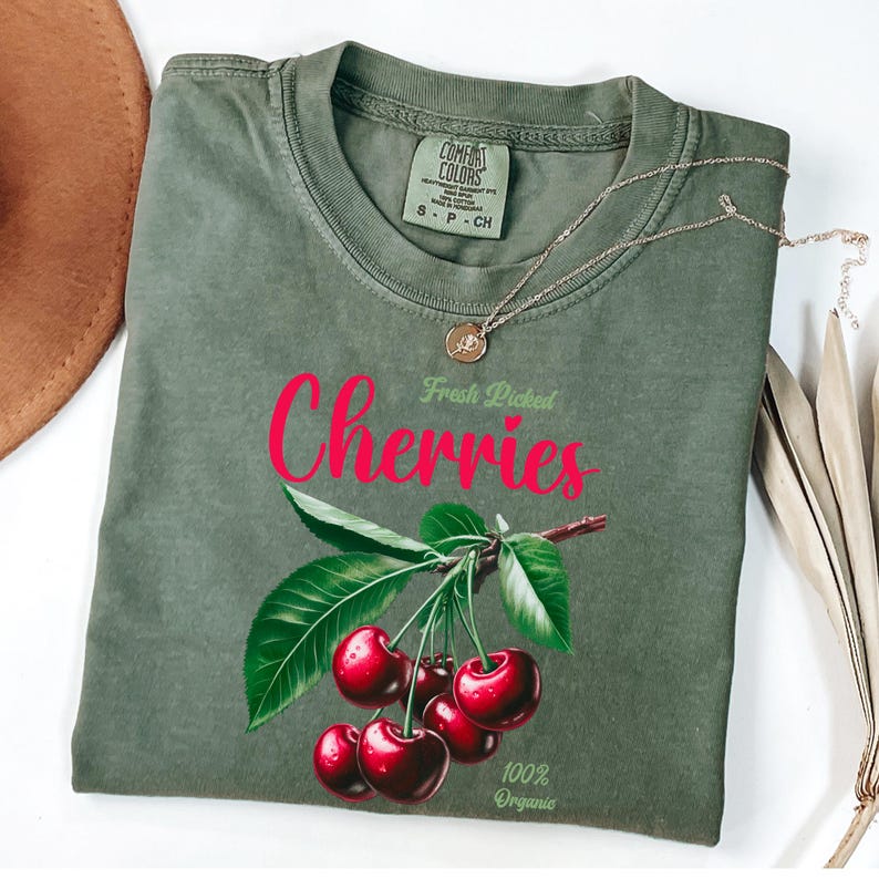 May include: A green t-shirt with a graphic of cherries on a branch with green leaves. The text "Fresh Picked Cherries" is above the cherries and the text "100% Organic" is below the cherries.