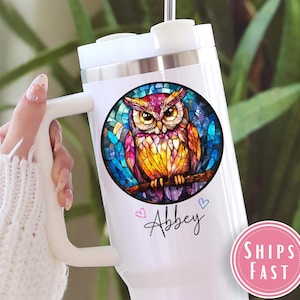 May include: White travel mug with a colorful owl design. The owl is perched on a branch within a stained-glass style circle. The name "Abbey" is written in cursive below the owl, with two small hearts.