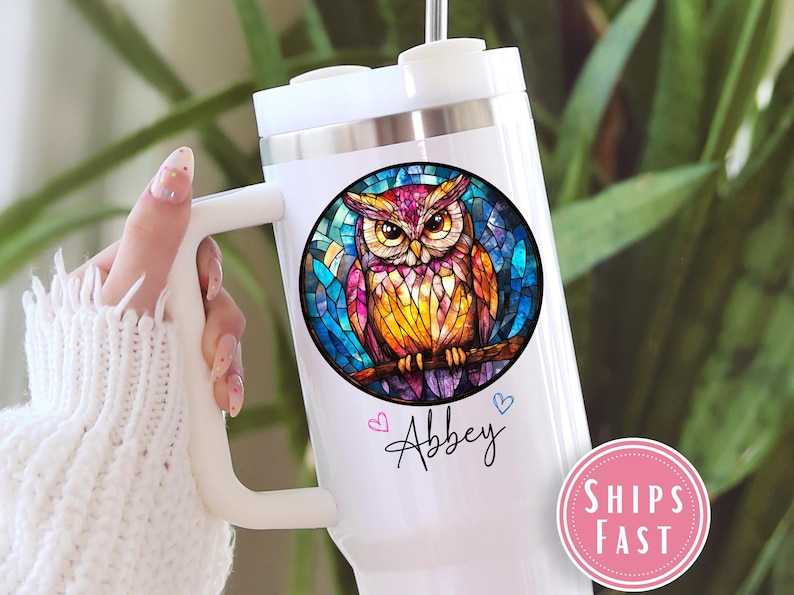 May include: White travel mug with a colorful owl design. The owl is perched on a branch within a stained-glass style circle. The name "Abbey" is written in cursive below the owl, with two small hearts.