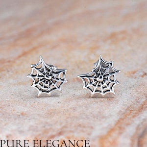 May include: A pair of silver-colored spiderweb stud earrings. Each earring features a detailed web design with a heart-shaped center. The earrings are displayed on a textured, neutral-toned surface. The text "PURE ELEGANCE" is at the bottom.