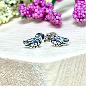 May include: A pair of small, silver-colored stud earrings shaped like detailed wings. The earrings are displayed on a light-colored wooden surface, with blurred purple flowers in the background. The earrings have a textured, feathered design.