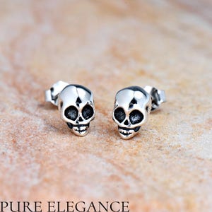 May include: A pair of silver skull stud earrings. The skulls are detailed with black eye sockets and a small mouth.