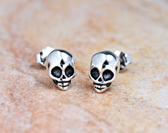 Könnte beinhalten: Ein Paar silberne Totenkopf-Ohrstecker. Die Totenköpfe sind mit schwarzen Augenhöhlen und einem kleinen Mund detailliert.