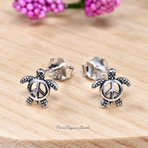 May include: A pair of sterling silver stud earrings shaped like sea turtles. The shell of each turtle features a cutout peace sign symbol. The earrings have a polished finish with darkened recessed areas to highlight the textured details of the flippers.