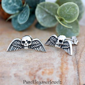 May include: A pair of silver skull earrings with wings. The skulls are detailed and the wings are spread out.