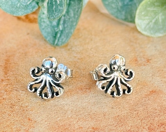 May include: A pair of silver-toned stud earrings shaped like small octopuses. Each earring features a detailed head with two prominent eyes and eight curled tentacles extending outward, finished with a metallic sheen and dark recessed accents.