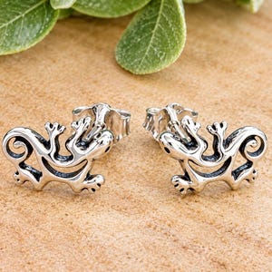 May include: A pair of silver stud earrings shaped like stylized lizards. Each earring features a polished metallic finish with darkened recessed areas to define the creature's body, limbs, and curled tail, complete with a standard post and backing.