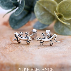 May include: A pair of silver-colored lizard stud earrings. The earrings are detailed with textured scales and small feet. The image is set against a blurred background of green foliage and a textured surface. The text "PURE ELEGANCE" is at the bottom.