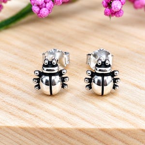 May include: A pair of polished silver stud earrings shaped like small beetles. Each earring features a rounded body with a central vertical line, a distinct head section, and six small legs extending from the sides. The studs include silver backings.