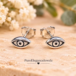 May include: A pair of sterling silver stud earrings shaped like eyes, featuring a central pupil and dark oxidized detailing. Each earring is stamped with 925 on the back post. The text PureEleganceJewelz is printed below the earrings.