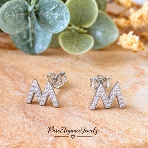 May include: A pair of silver stud earrings shaped like the letter M, encrusted with small clear stones. The back of the earring post is clearly marked with 925, indicating sterling silver. The text PureEleganceJewelz is visible below.