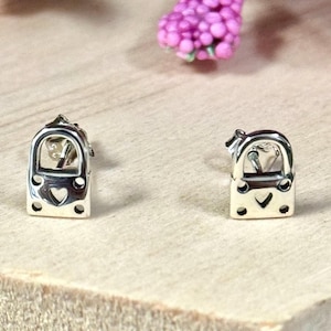 May include: A pair of silver stud earrings shaped like miniature padlocks. Each earring features a polished metallic finish with a small heart-shaped cutout in the center and two tiny circular indentations at the base, secured with standard earring backs.