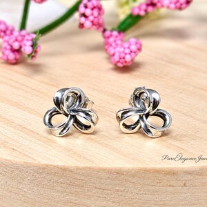 May include: A pair of silver stud earrings featuring a three-looped knot design with polished surfaces and darkened recessed areas. The earrings are positioned side by side, displaying a symmetrical, rounded, and interconnected metal structure.