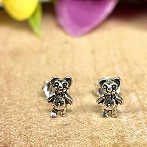 May include: A pair of silver-colored bear stud earrings. Each earring features a small, detailed bear figure with arms, legs, and facial features. The earrings are set against a neutral background, highlighting the jewelry's design.