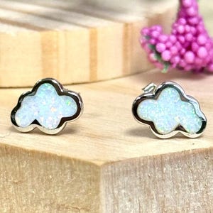 May include: A pair of cloud-shaped stud earrings featuring a polished silver-tone metal border and a white iridescent opal-like inlay with subtle flecks of blue and green color throughout the surface of each individual cloud-shaped earring piece.