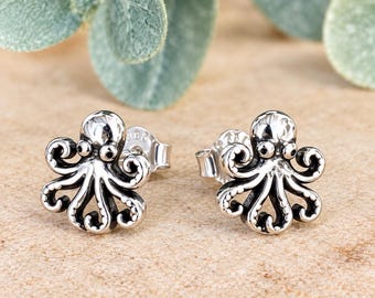 Può includere: Un paio di orecchini a lobo a forma di polpo color argento, caratterizzati da un design dettagliato con tentacoli arricciati e occhi sporgenti. Il metallo presenta una finitura lucida con zone scure per evidenziare la texture del polpo.