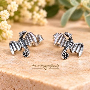 May include: A pair of silver-toned stud earrings shaped like dragons with spread wings and curled tails. The metal features oxidized detailing to highlight the textured scales and wing structure. The text PureEleganceJewelz with a heart icon is visible.