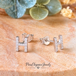 May include: A pair of silver-toned stud earrings shaped like the letter H, featuring small clear stones embedded along the surface. The back of the earring post is stamped with 925, indicating sterling silver composition.