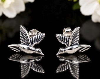 May include: A pair of sterling silver stud earrings shaped like hummingbirds in flight. The metal has a polished finish with darkened recessed areas to highlight the wing feathers and body details. Each earring features a standard post back.