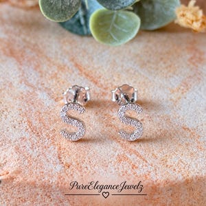 May include: A pair of silver stud earrings shaped like the letter S, each featuring a pavé setting of small clear stones. The earrings have a post back closure and are displayed on a textured surface with the text PureEleganceJewelz.