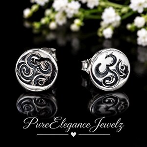 May include: A pair of round silver stud earrings featuring a raised, polished Om symbol set against a recessed, oxidized black background. The earrings have a smooth, circular border and are displayed with their reflections on a black surface.