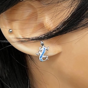 May include: A silver-toned stud earring shaped like a lizard, featuring a vibrant blue inlay along its back. The lizard design is detailed with four legs and a curved tail, providing a distinct contrast between the metallic silver finish and the blue.