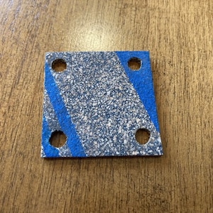 Puede incluir: Una junta de corcho cuadrada con cuatro agujeros y rayas pintadas de azul. La junta está hecha de un material rugoso y texturizado.