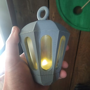 May include: A small grey lantern with a yellow light inside. The lantern has a loop at the top for hanging.