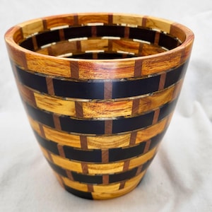 May include: A tapered wooden bowl featuring a segmented brick-like pattern. The design alternates between horizontal light brown wood segments and dark black rectangular pieces, joined by small vertical brown wood connectors throughout.