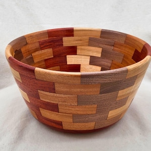 Può includere: Una ciotola rotonda in legno costruita con piccoli blocchi rettangolari di varie essenze legnose. La ciotola presenta un motivo a mosaico con tonalità di legno alternate in beige chiaro, marrone medio, marrone scuro e marrone rossiccio.